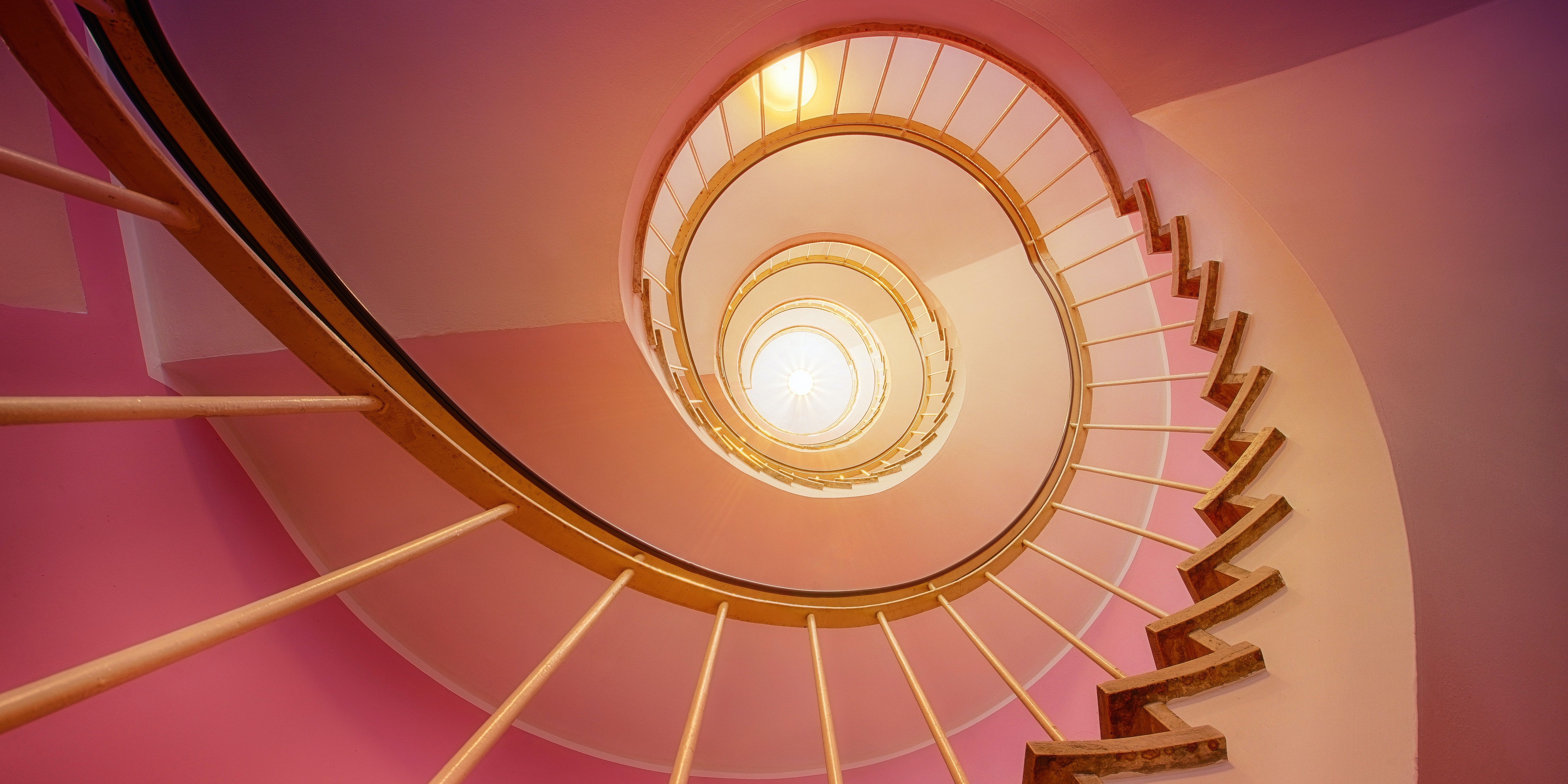View looking up toward a skylight through many floors of a spiral staircase, in warm pink and yellow hues